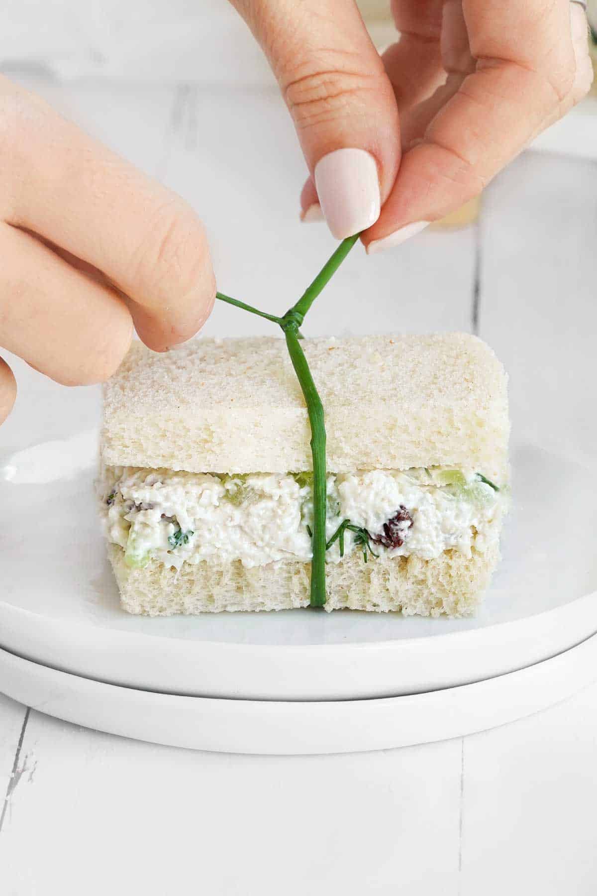 Hands tying a chive around a sandwich on a white plate.