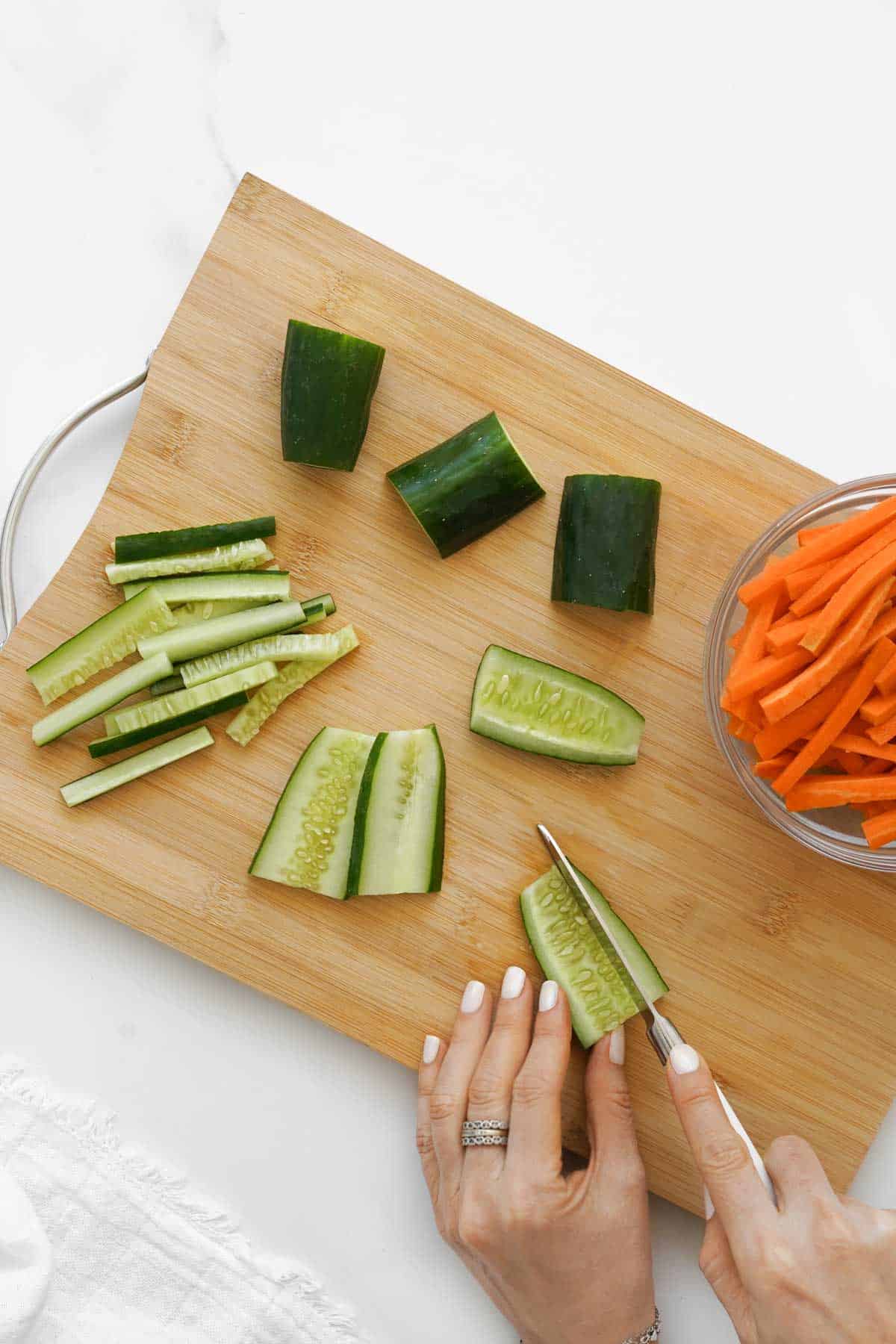 A person slicing cucumber on a wooden cutting board, next to a bowl of carrot sticks.