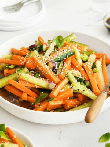 A bowl of carrot and cucumber salad with sesame seeds, accompanied by a side of dressing.