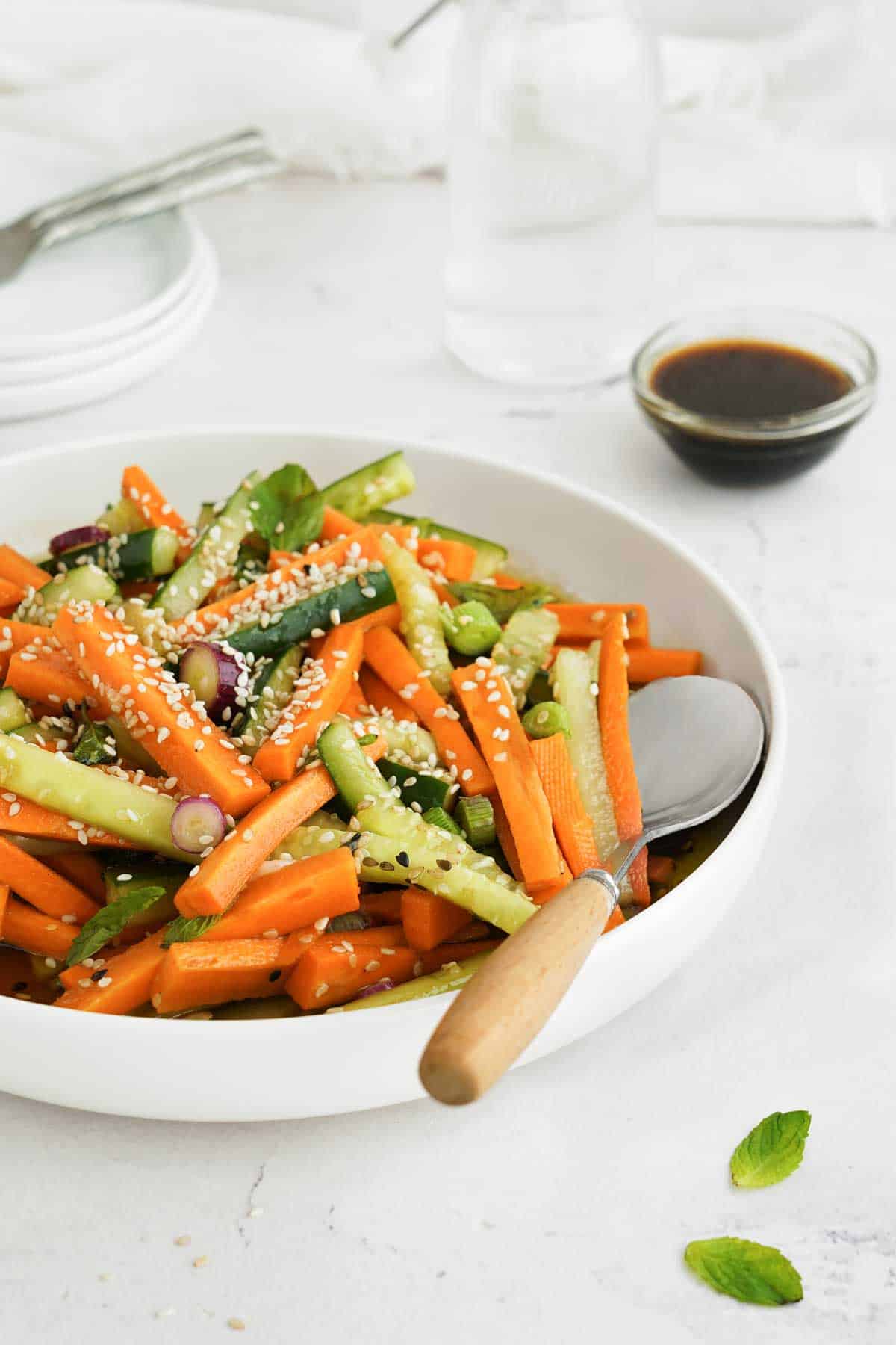 A bowl of carrot and cucumber salad with sesame seeds, accompanied by a side of dressing.