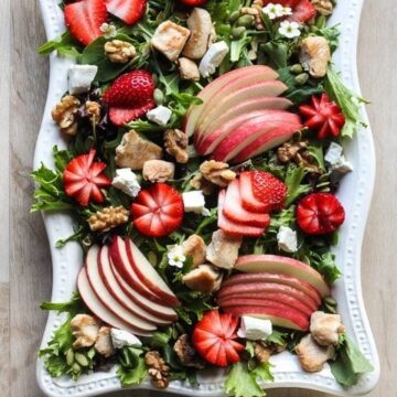 Fresh mixed greens salad with strawberries, apple slices, feta cheese, walnuts, and croutons, served with a side of dressing.