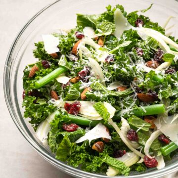 A glass bowl containing a kale salad with shaved parmesan cheese, dried cranberries, and almonds.