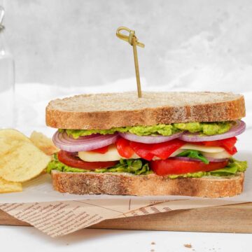 A vegetable Mediterranean sandwich is sitting on a cutting board next to chips.