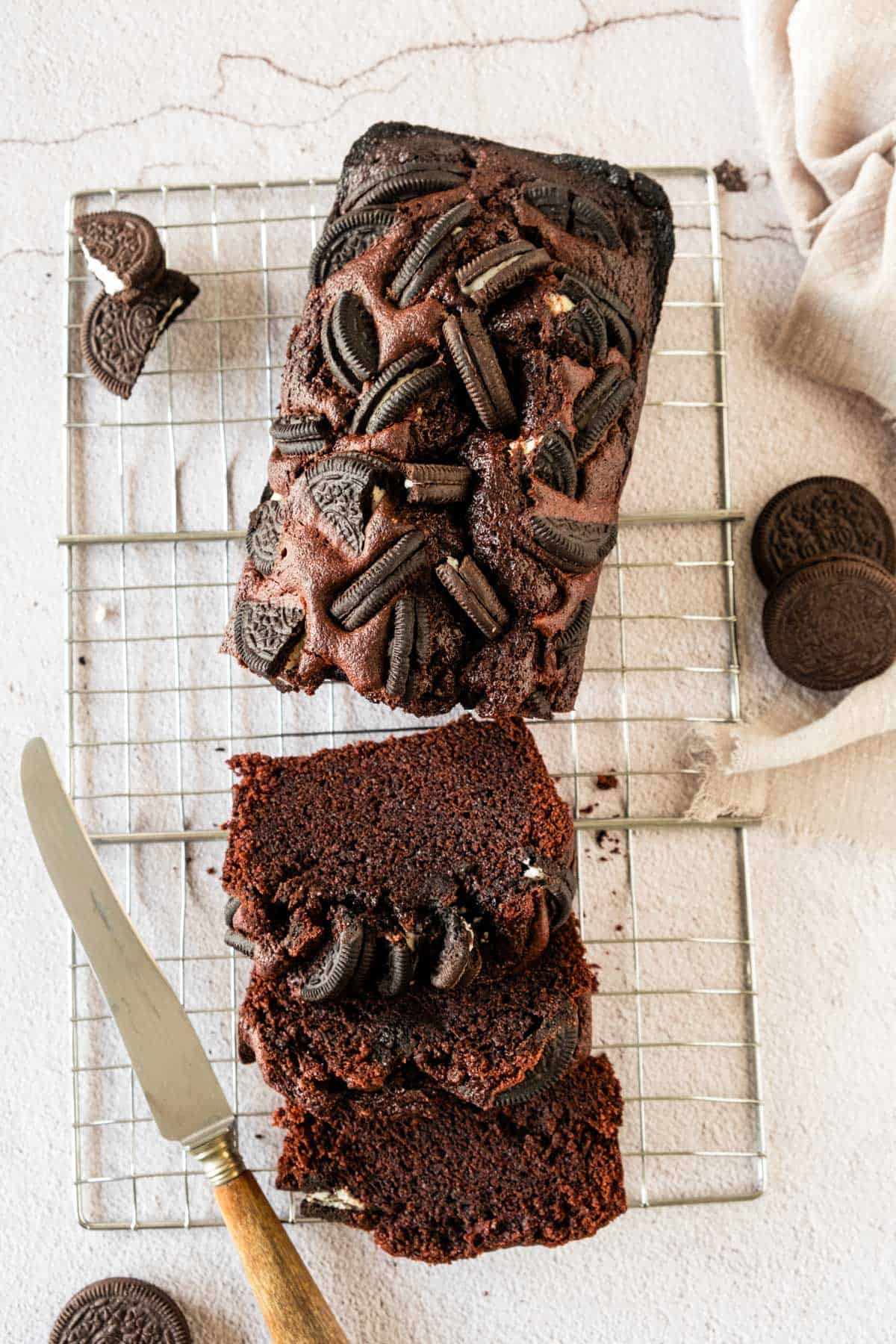 Chocolate loaf cake topped with cream and cookie pieces, with one slice partly cut, on a wire rack with cookies nearby.