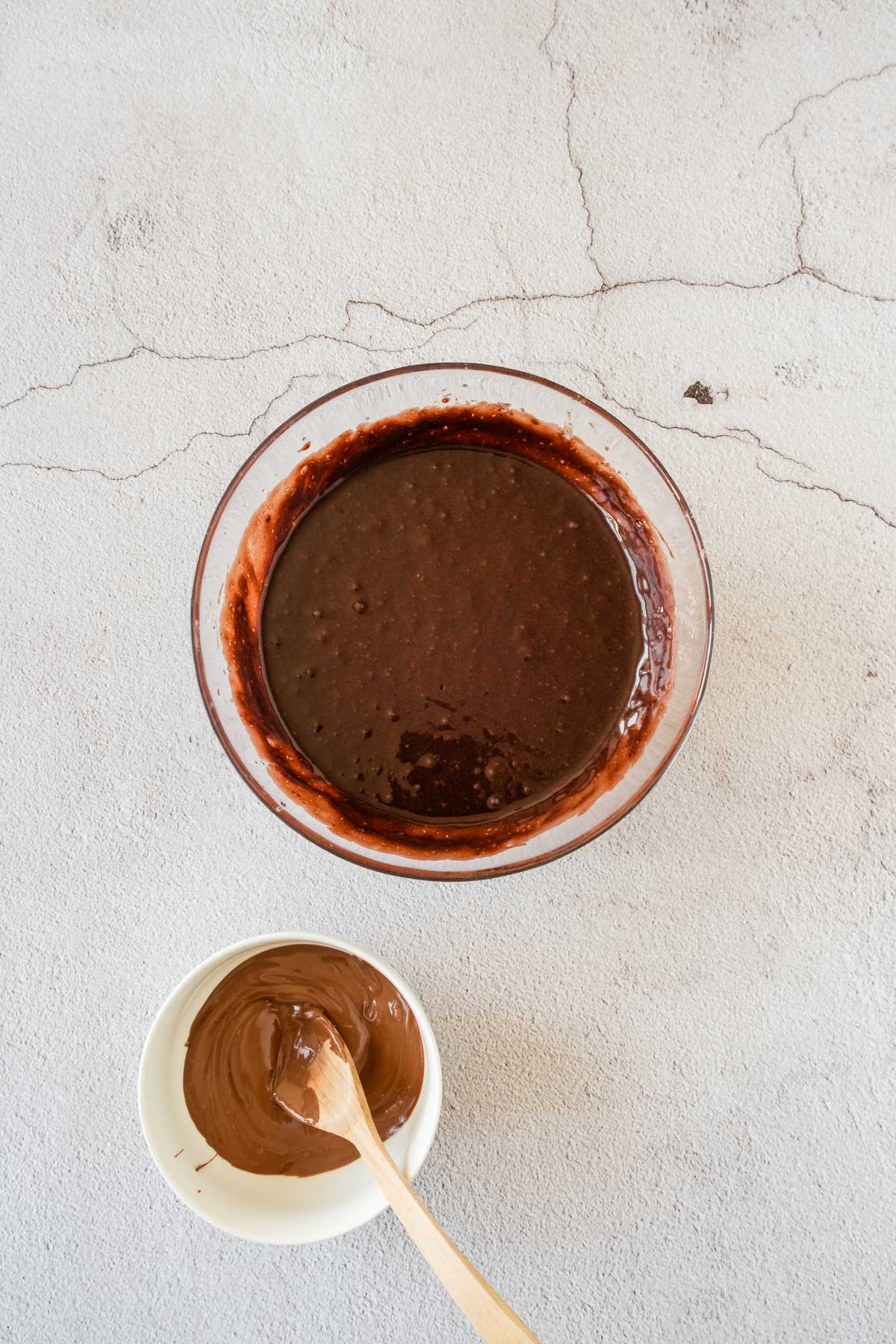 A bowl of mixed chocolate batter alongside a smaller bowl with melted chocolate and a wooden spoon, on a textured white surface.
