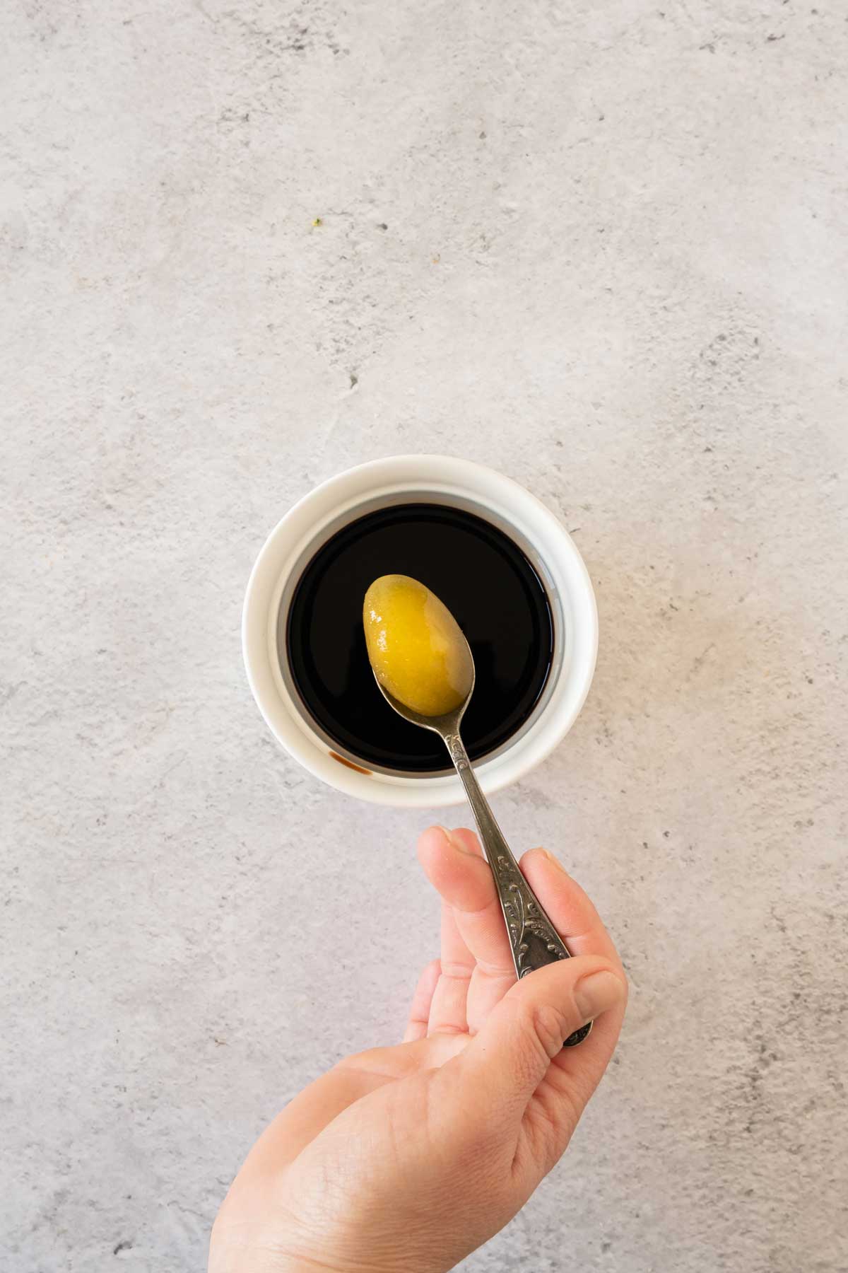 A hand holding a spoon with honey in it over a bowl of Balsamic Vinegar.