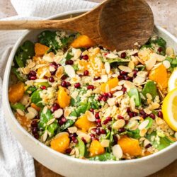 A colorful bowl of salad with quinoa, spinach, mandarin oranges, pomegranate seeds, and almond slices, with a wooden spoon on the side.