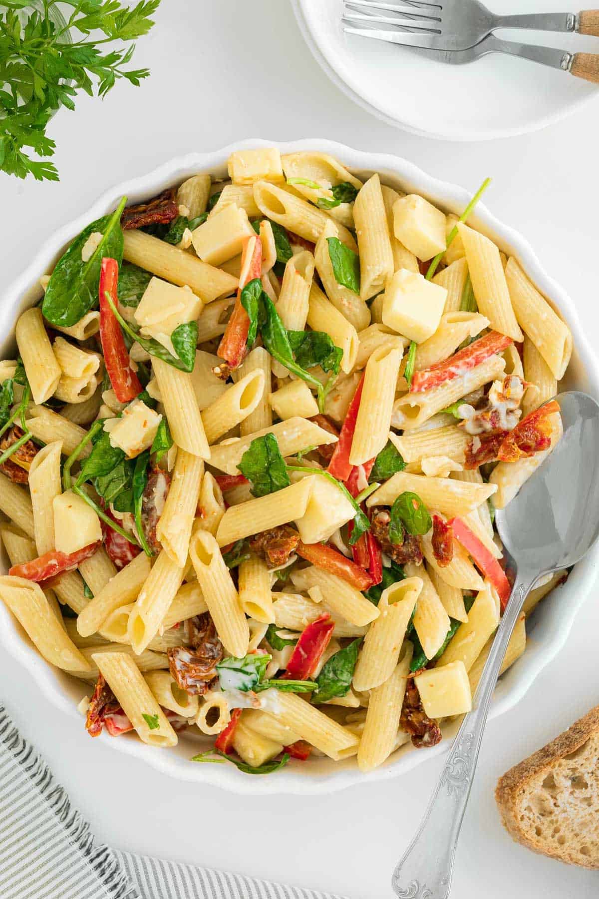 A bowl of penne pasta salad with spinach, sun-dried tomatoes, and cheese on a table with utensils and bread.