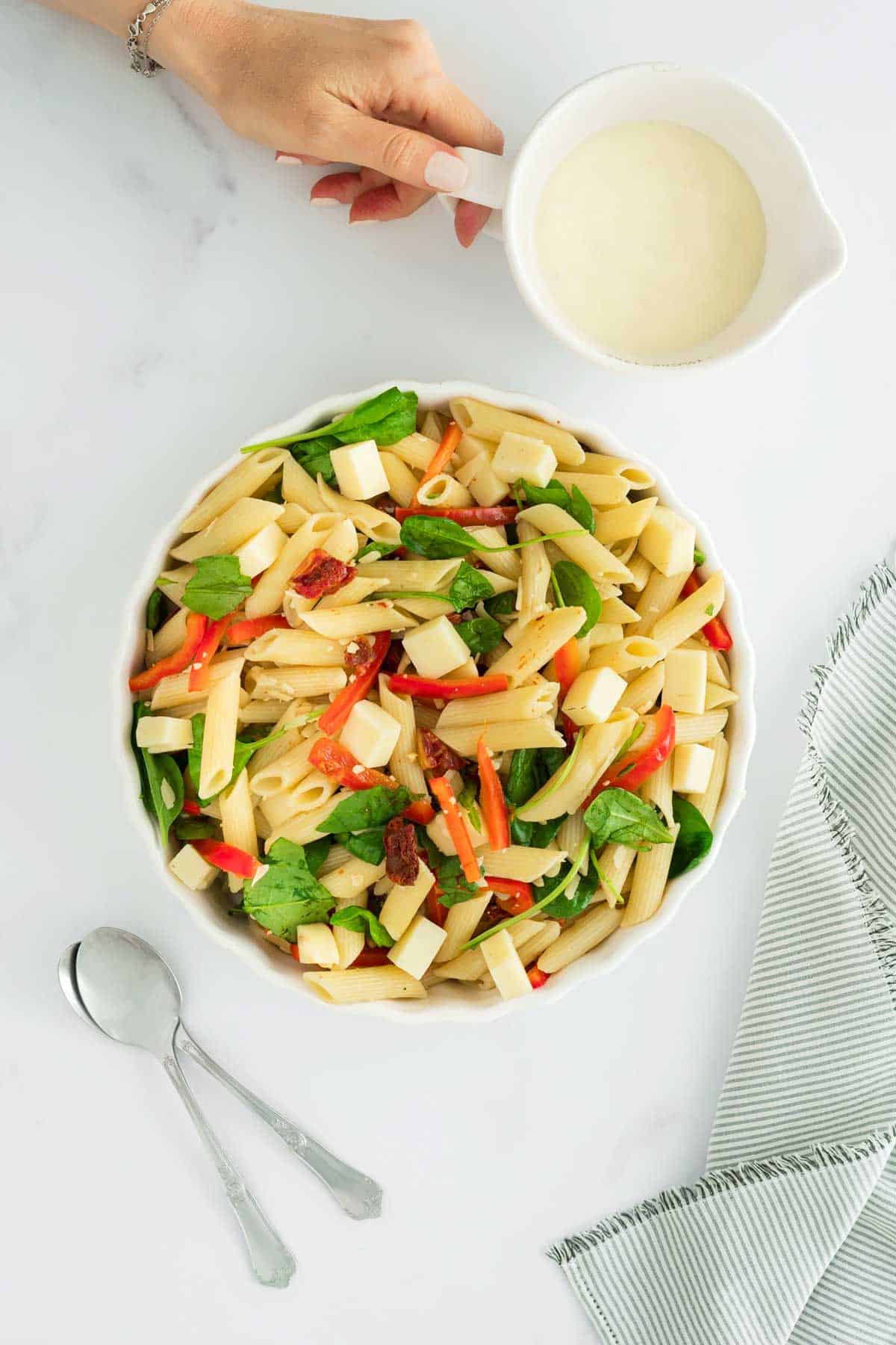 A bowl of pasta salad with vegetables and a hand holding a dressing jar.
