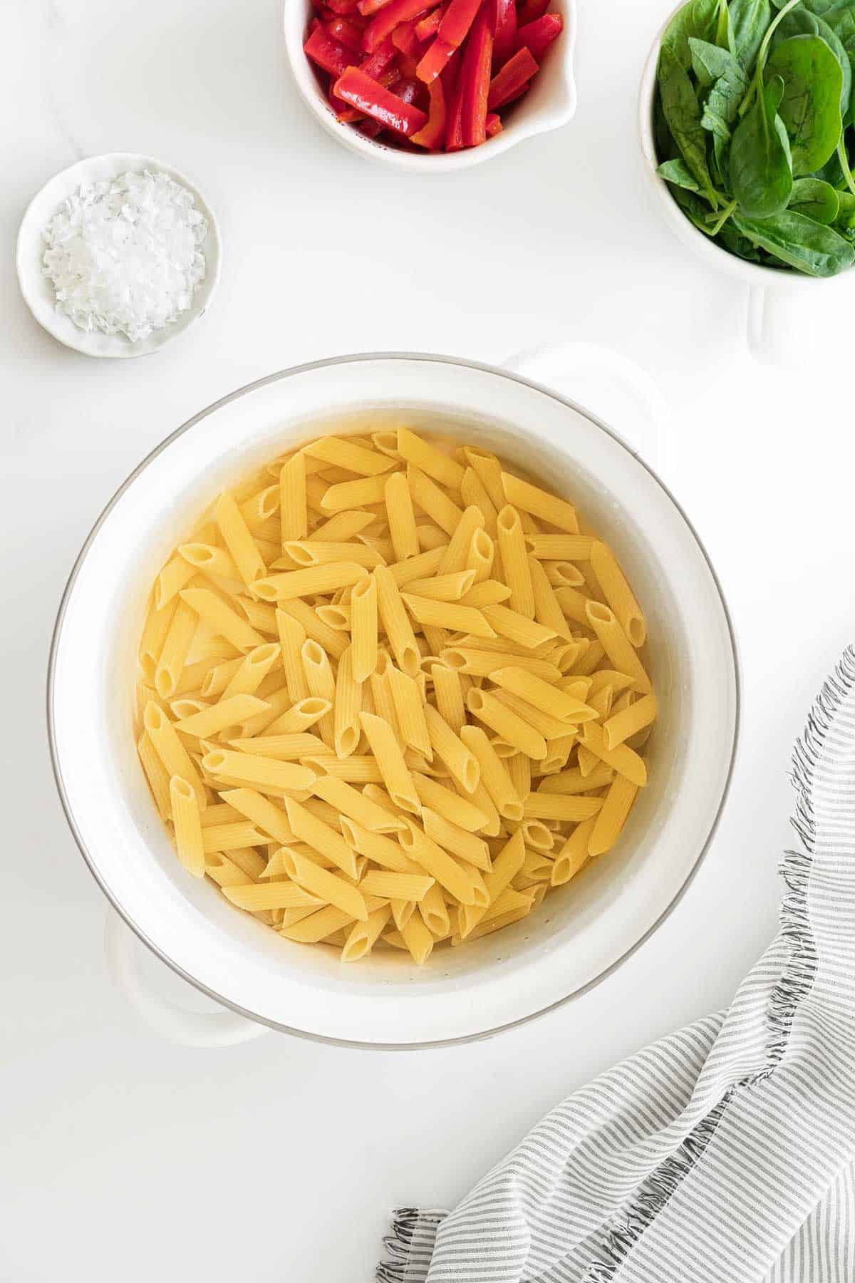 A bowl of raw penne pasta with ingredients like spinach and red pepper on the side, ready for cooking.