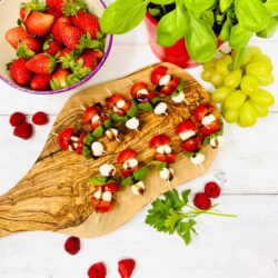 A selection of fresh caprese salad skewers with tomatoes, basil, and mozzarella cheese, accompanied by bowls of strawberries and grapes on a wooden table.