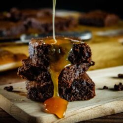 Caramel being poured onto a stack of brownies on a wooden board.