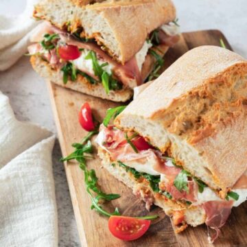 A sandwich with ham, cheese, tomatoes, and greens on a wooden cutting board.
