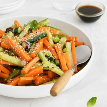 A bowl of vegetable salad with carrot sticks, cucumber slices, green peas, and a sprinkling of sesame seeds, accompanied by a side of soy sauce dressing.