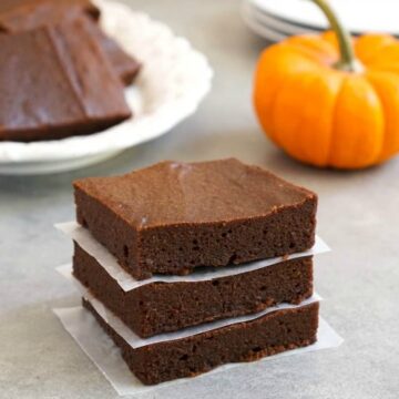 Stack of three chocolate brownies with more on a plate in the background, accompanied by a small orange pumpkin on a kitchen counter.