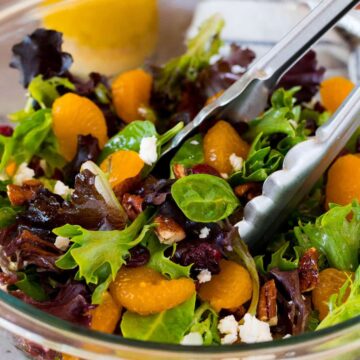 A person tossing a mixed greens salad with mandarin oranges, feta cheese, and nuts in a glass bowl.