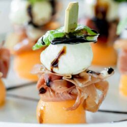 Elegant appetizer arrangement featuring melon topped with prosciutto, mozzarella, basil, and balsamic glaze.