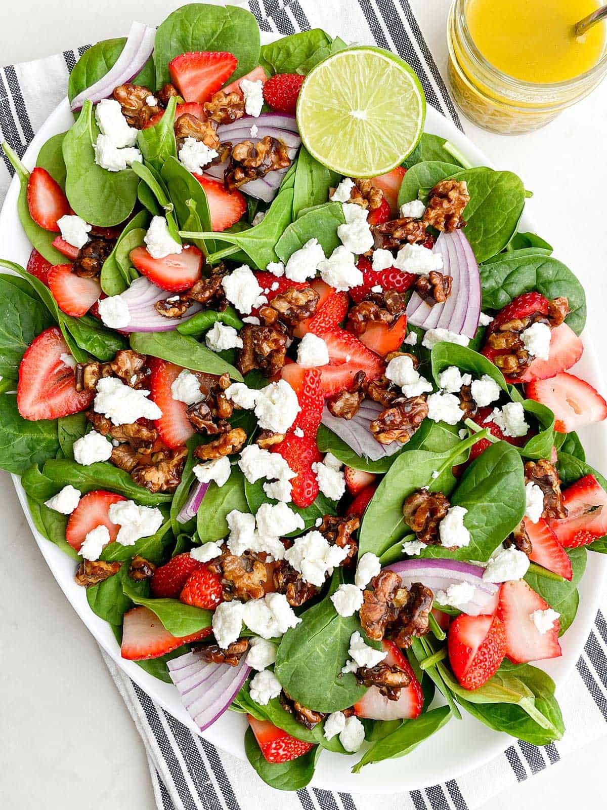A fresh spinach salad with strawberries, walnuts, and goat cheese, served with a lime wedge and a glass of juice on the side.