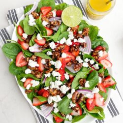A fresh spinach salad with strawberries, walnuts, and goat cheese, served with a lime wedge and a glass of juice on the side.