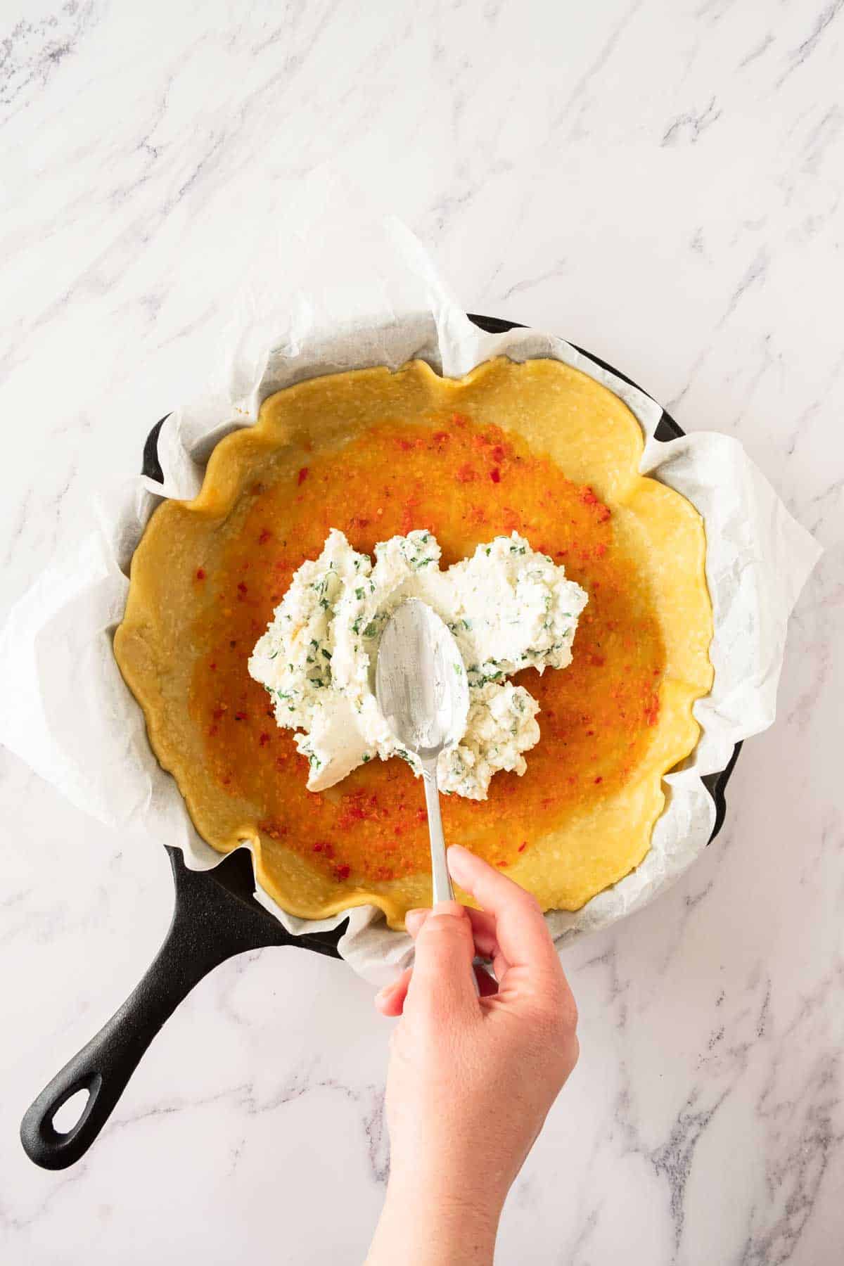 A hand spooning cottage cheese onto a galette pastry base in a cast iron pan.