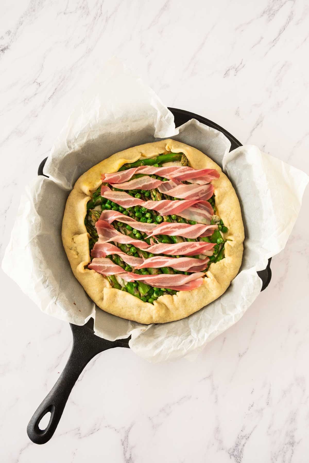 An asparagus galette in a cast iron pan with peas topped with bacon strips during the cooking process.