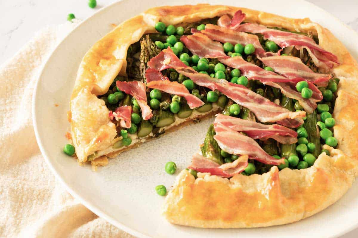 An asparagus galette topped with fresh peas and bacon. A slice has been cut from the pie.