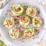 Cucumber roll-ups filled with cream cheese, smoked salmon, and herbs, garnished with chili flakes on a plate.