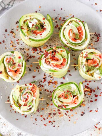 Cucumber roll-ups filled with cream cheese, smoked salmon, and herbs, garnished with chili flakes on a plate.
