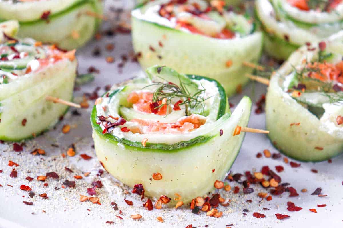 Cucumber roll ups filled with cream cheese, smoked salmon, and dill, garnished with red pepper flakes on a white plate.