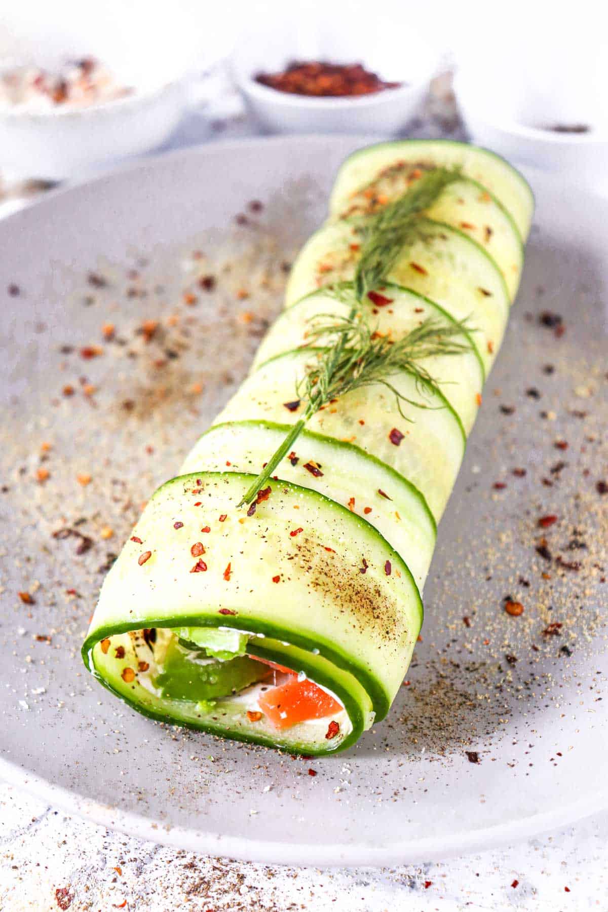 A cucumber roll filled with cream cheese and smoked salmon, garnished with dill and sprinkled with spices on a gray plate.