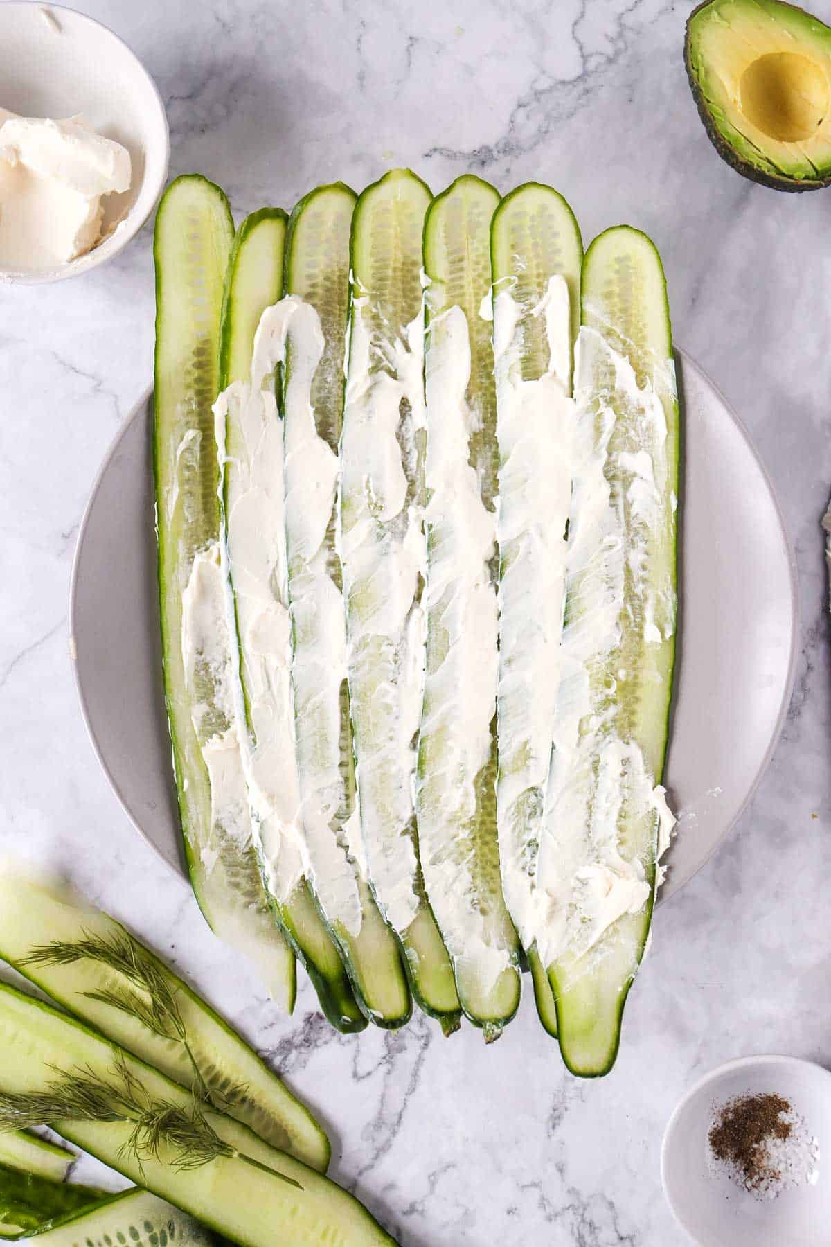 Thinly sliced cucumber lined up on a plate, topped with cream cheese, surrounded by ingredients like avocado, and seasonings.
