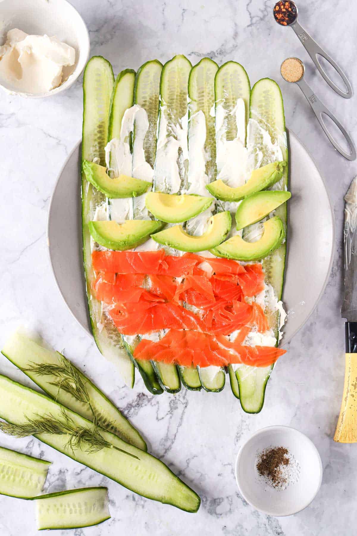 Cucumber slices, cream cheese, avocado, and smoked salmon, arranged on a plate with dill, seasoning, and cooking ingredients visible.