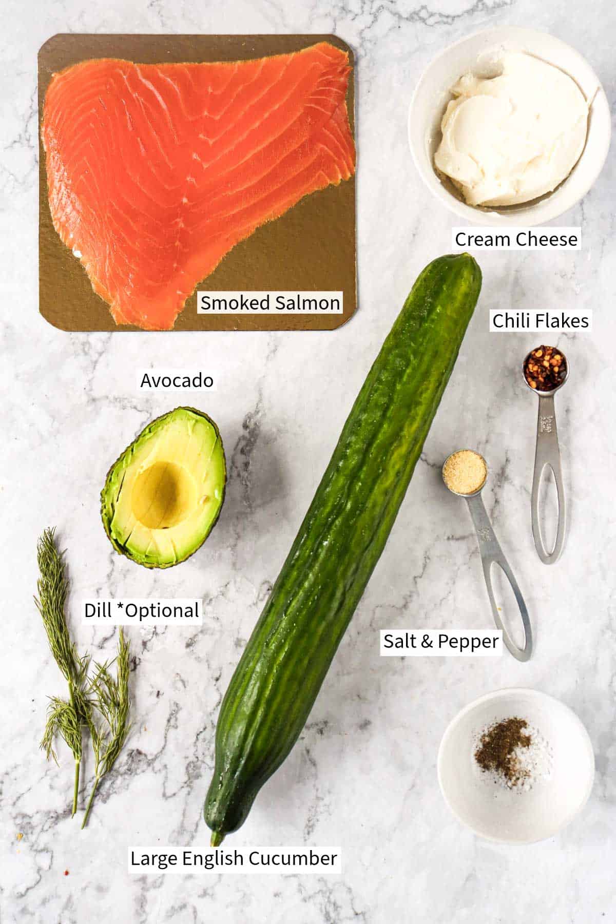 Ingredients for cucumber roll ups displayed on a marble table including smoked salmon, cream cheese, an avocado, cucumber, dill, chili flakes, and seasonings.