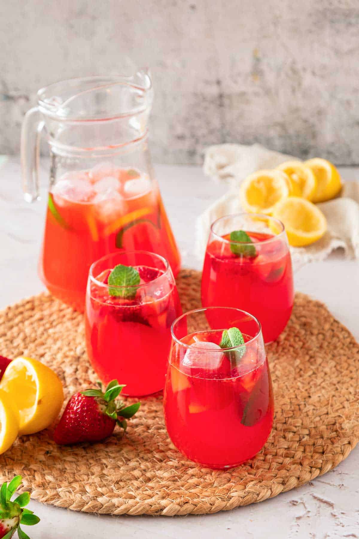 A jug and glasses of strawberry lemonade on a table surrounded by squeezed lemons and strawberries.