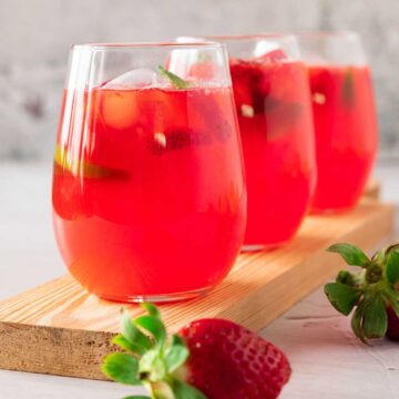 Three glasses of pink strawberry lemonade in a line with squeezed lemons and strawberries scattered around.
