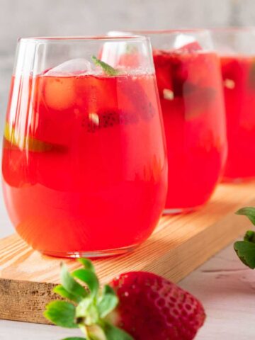 Three glasses of pink strawberry lemonade in a line with squeezed lemons and strawberries scattered around.