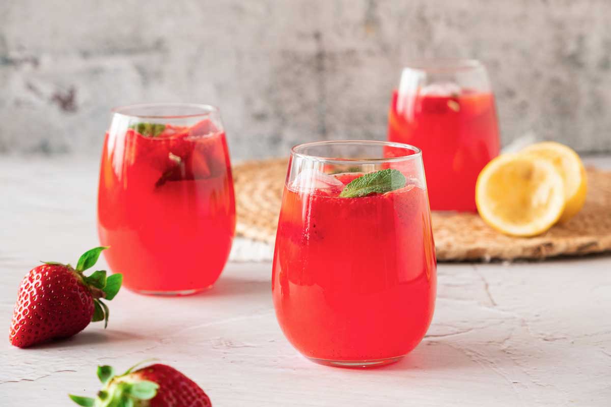 Three glasses of pink strawberry lemonade with squeezed lemons and strawberries scattered around.