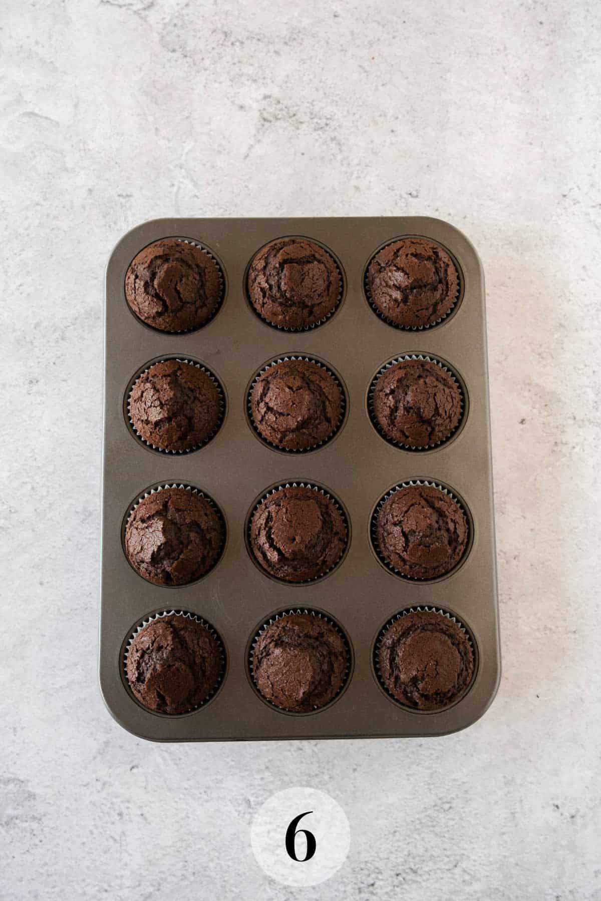 A dozen freshly baked chocolate cupcakes in a brown muffin tray, arranged in four rows on a light grey surface.