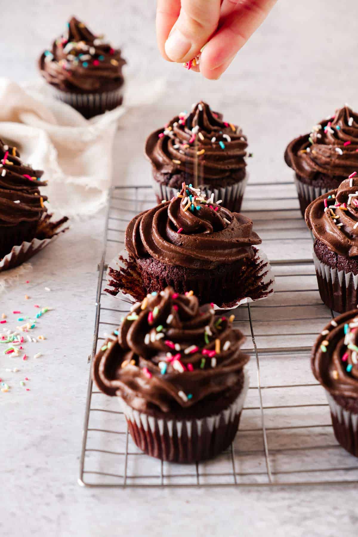 A hand sprinkling colorful sprinkles on chocolate cupcakes with swirls of chocolate frosting arranged on a wire cooling rack.