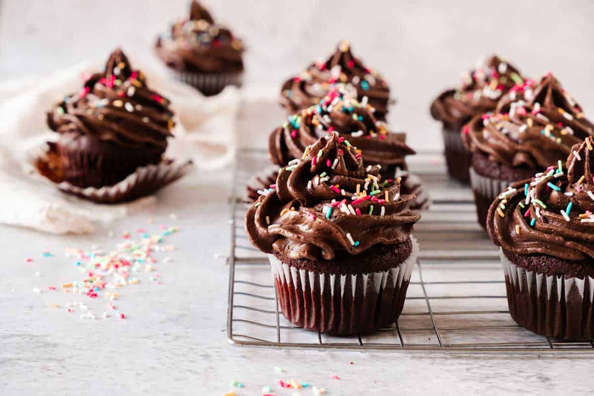 Double chocolate cupcakes with chocolate frosting and sprinkles on a cooling rack against a light background.