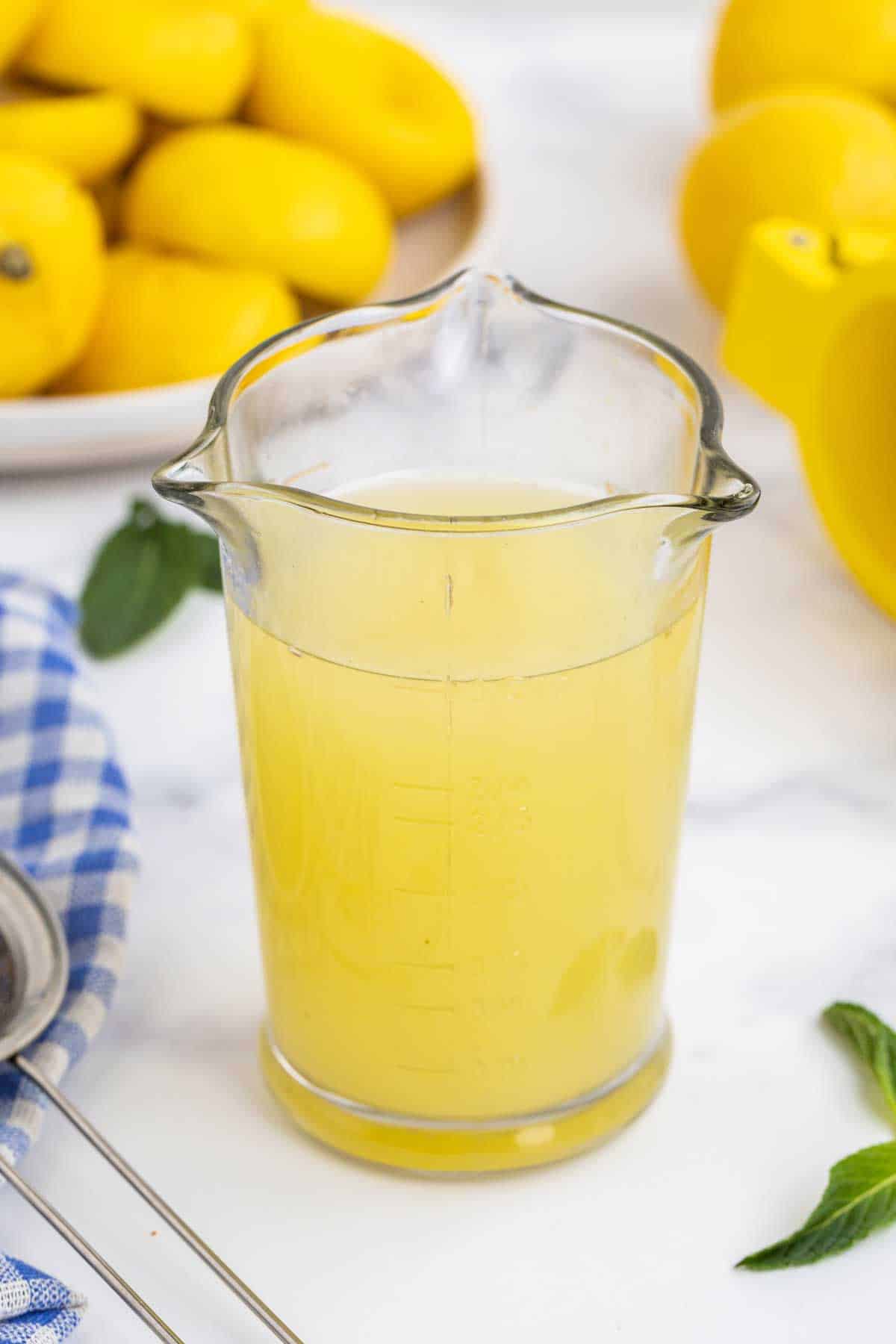 A glass pitcher filled with lemon juice, with lemons and a blue checkered cloth in the background.