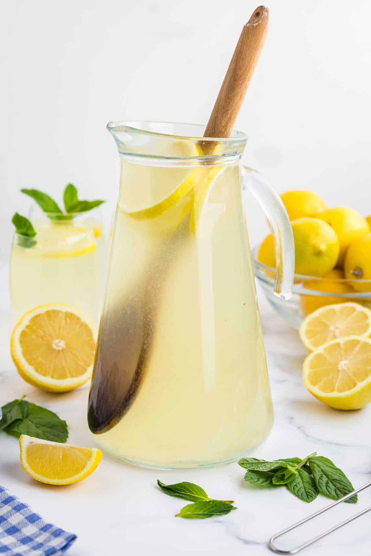 A pitcher of lemonade with lemon slices and a wooden stirrer, accompanied by a bowl of lemons and mint leaves.