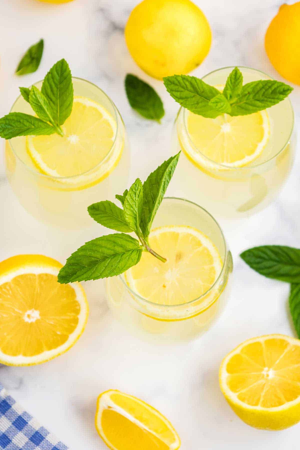 Three glasses of lemonade garnished with lemon slices and mint leaves on a marble surface, with whole and sliced lemons nearby.