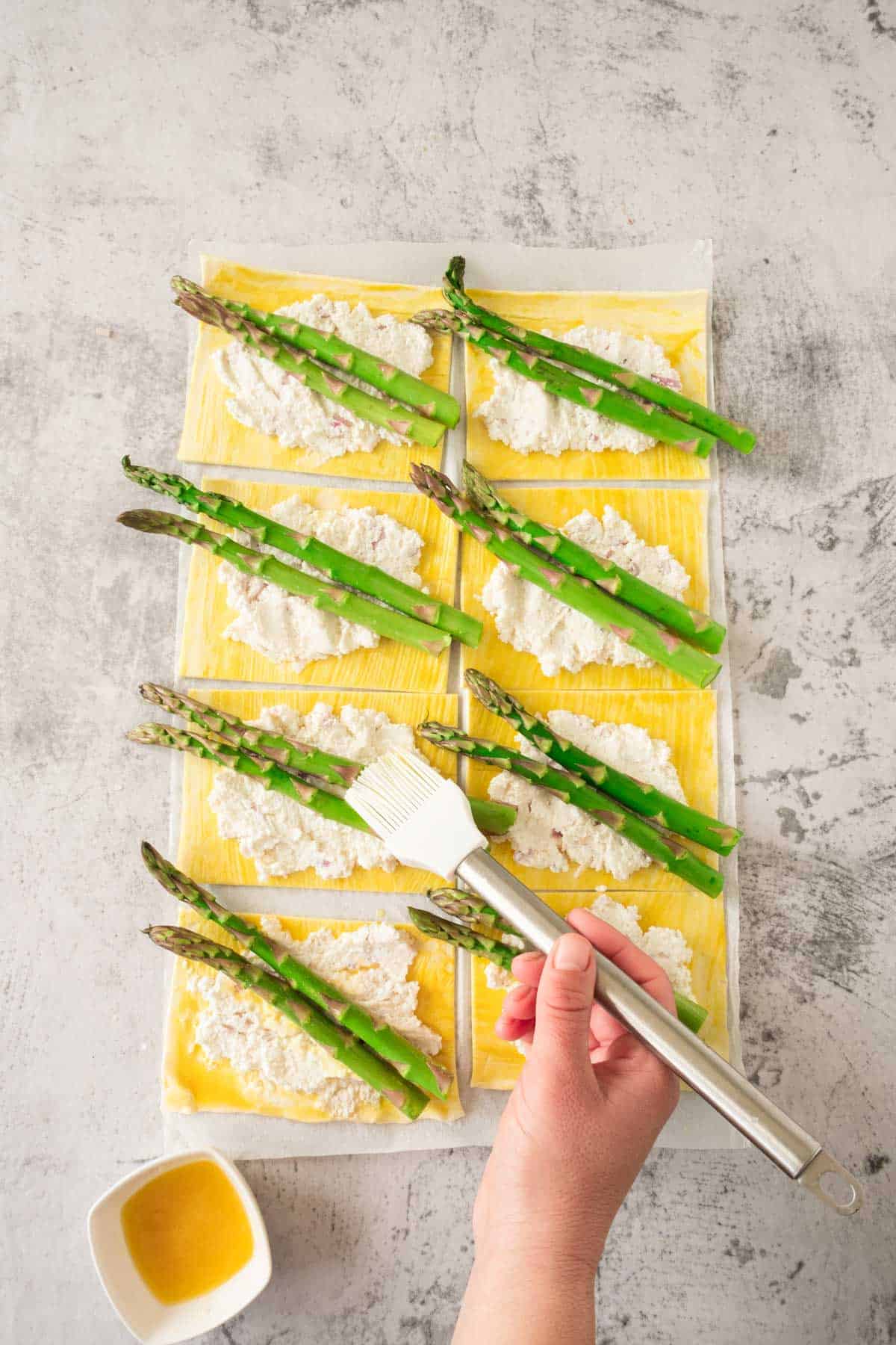 Preparing asparagus-topped puff pastry sheets with ricotta cheese, with a hand brushing on butter before baking.
