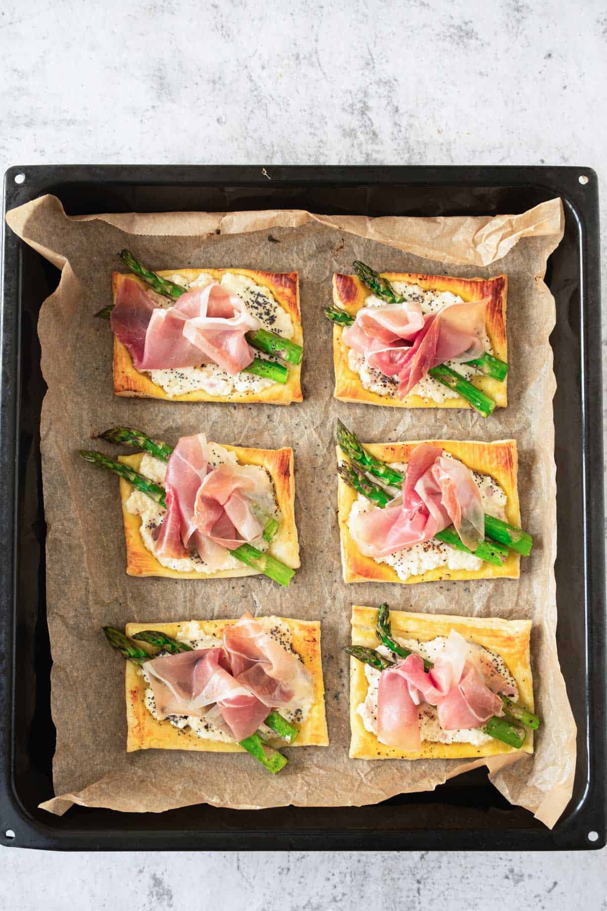 A tray of freshly baked puff pastry tarts with asparagus and prosciutto on a parchment paper.