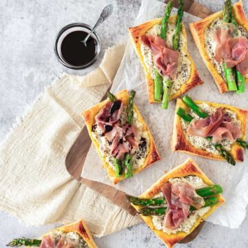 Slices of asparagus and prosciutto tart on a wooden board with a jar of balsamic sauce.