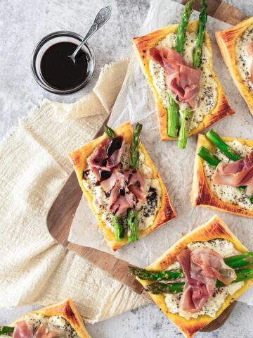 Slices of asparagus and prosciutto tart on a wooden board with a jar of balsamic sauce.