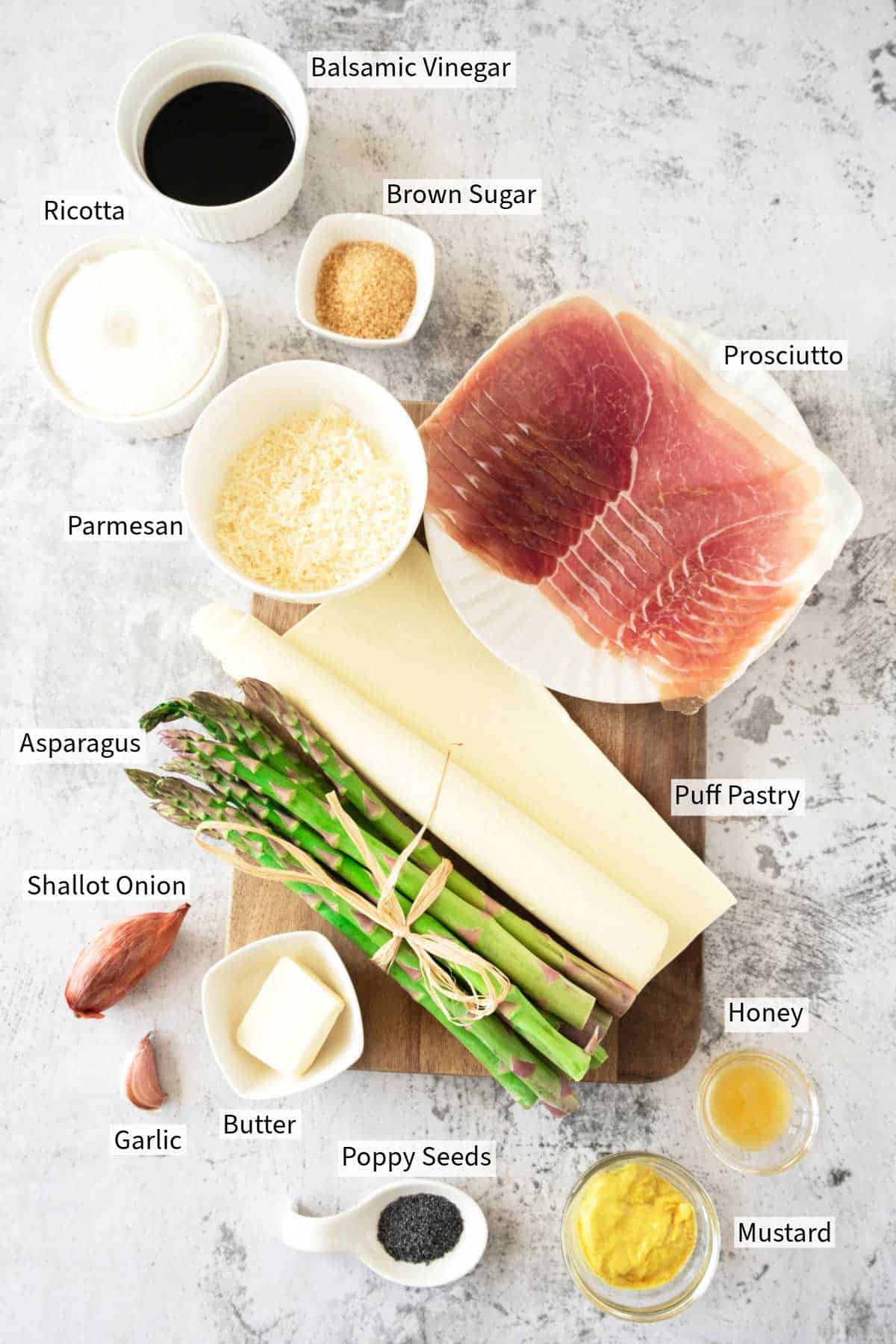 Ingredients for a asparagus tart recipe laid out on a table, including puff pastry, prosciutto, asparagus, and various condiments and cheeses.