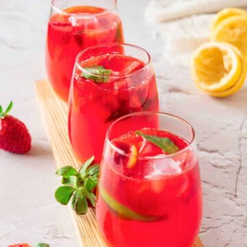 Three glasses of strawberry lemonade with mint leaves and ice on a wooden board, surrounded by fresh strawberries and lemon slices.