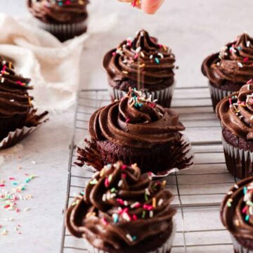 A person sprinkles colorful toppings on a row of chocolate cupcakes with swirls of rich frosting on a cooling rack.